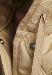 Beige vuorattu takki, jossa on tekstuuri, näkyvät ompeleet ja nepparikiinnitys. Vuori tuo esiin hienovaraisen kiillon.