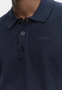 Marinblå polo skjorta med stickad textur, med en klassisk krage, tre knappar och broderad branding i en mörkare nyans.