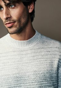 Suéter de punto gris claro con un patrón texturizado, que presenta un cuello acanalado y una tela suave y transpirable. Primer plano del cuello y el área del hombro.