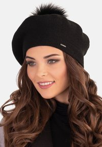 Fekete kötött beret, puha műszőrme pom-pommal a tetején, diszkrét fémből készült címkével, laza, omlós szabással.