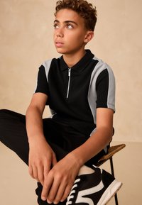 Černo-šedé polo tričko s krátkými rukávy na zip, doplněné o černé kalhoty a černé tenisky. Kudrnaté vlasy, sedící pozice.