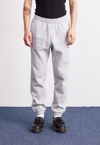 Grå bomullsmjukisbyxor med resår i midjan och avsmalnande muddar, med en liten röd- och vit logotyp på vänster sida. Svarta skor med detaljer.