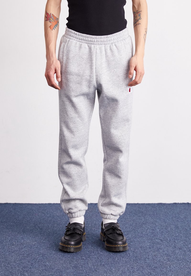 Grå bomullsmjukisbyxor med resår i midjan och avsmalnande muddar, med en liten röd- och vit logotyp på vänster sida. Svarta skor med detaljer.