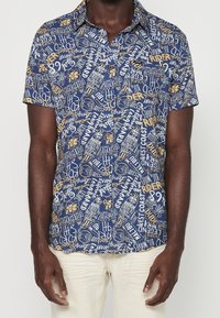 Homme portant une chemise bleu marine à manches courtes, boutonnée, avec un texte graphique blanc et jaune et des motifs floraux, associée à un pantalon beige clair.