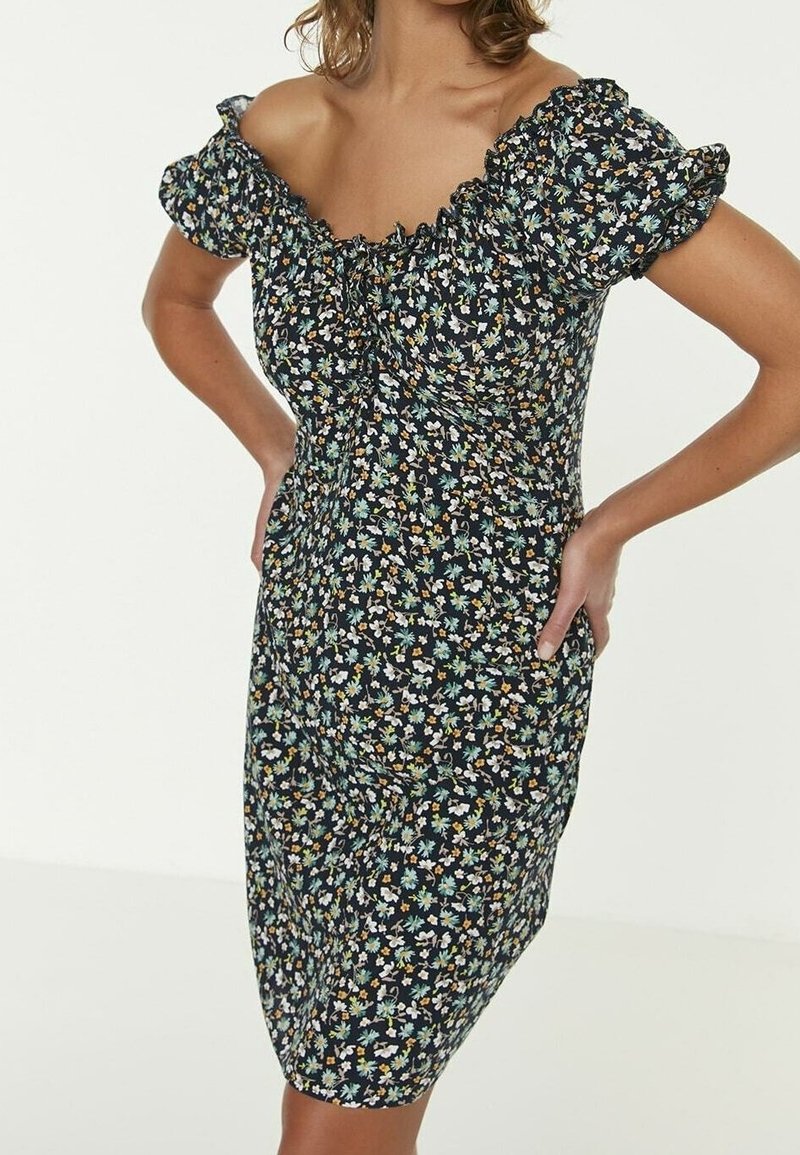 Femme portant une robe noire ajustée avec un petit imprimé floral bleu, blanc et jaune, des manches bouffantes et un décolleté décolleté.