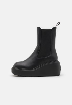Botte Chelsea en cuir noir avec semelle plateforme épaisse, panneaux latéraux élastiques et languette de tirage sur fond blanc uni.