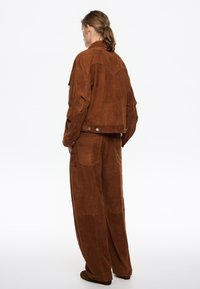 Bruine suede jas en wijde broek, met een kraag, rugpand detail en zakken. Gladde textuur, minimale hardware accenten.