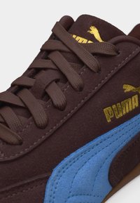 Καφέ σουέντ αθλητικό παπούτσι Puma με καφέ κορδόνια, μπλε πλαϊνή ρίγα και χρυσό λογότυπο Puma στη γλώσσα και στο πλάι.