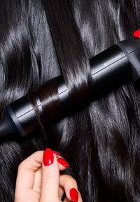 Un fer à coiffer noir positionné contre des cheveux lisses, foncés et brillants. Une mèche de cheveux est enroulée autour du fer. Des ongles manucurés rouges sont visibles.
