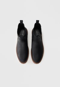 Par de botas de cuero negro hasta el tobillo con suelas marrones, lengüetas y paneles laterales de elastano sobre un fondo claro.