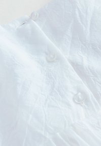 Hvid tekstureret stof med krøllet overflade; indeholder to hvide knapper. Designet er skåret i en let vinkel, hvilket fremhæver materialets blødhed.