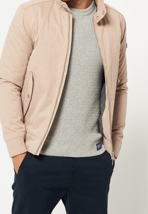 Homme portant une veste décontractée beige sur un pull en tricot gris clair et un pantalon bleu marine foncé, mains dans les poches, debout devant un fond uni.