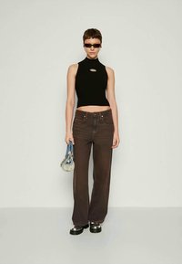Mulher vestindo um top cropped preto sem mangas, jeans largos castanho-escuros, sapatos pretos robustos, óculos de sol, segurando uma pequena bolsa de denim contra um fundo liso.