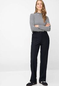 Gestreept shirt met lange mouwen in zwart en wit, gecombineerd met donkergekleurde, hooggetailleerde broek met taillebandlussen en zakken, gedragen met donkere schoenen.