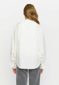 Weiße Baumwollbluse mit Ballonärmeln, die kunstvoll bestickte Spitzenakzente an den Manschetten und einen leicht gerüschten Halsausschnitt aufweist.