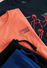 To sammenfoldede grafiske T-shirts, en koralfarvet med teksten "picky bits supper club" og en sort med rød stregtegning, samt to par bukser med snøre i sort og blå.