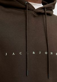 Hanorac maro cu cordon și accente metalice argintii. Prezintă un logo cu textul "JACK & JONES" în litere gri deschis pe față.