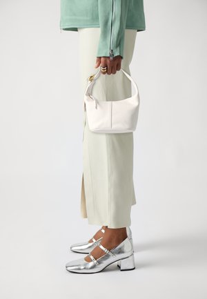 White leather handbag with a curved shape, held by a person. The outfit features light green fabric, complementing silver block-heeled shoes.