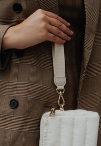 Sac matelassé blanc avec quincaillerie dorée, attaché à une bandoulière beige étiquetée "MARLY," tenu devant un fond en tissu écossais marron.