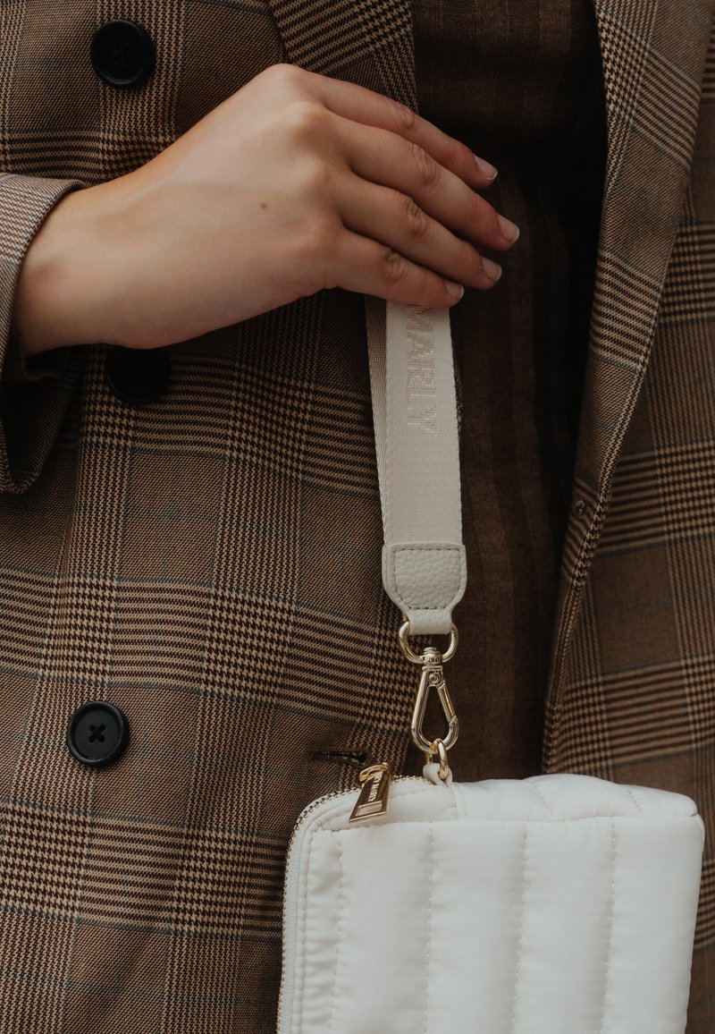 Sac matelassé blanc avec quincaillerie dorée, attaché à une bandoulière beige étiquetée "MARLY," tenu devant un fond en tissu écossais marron.
