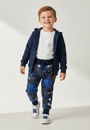 Uśmiechnięty młody chłopiec idący, ubrany w granatową bluzę z kapturem na zamek, białą koszulkę, spodnie z nadrukiem galaktyki oraz białe trampki z niebieskimi sznurowadłami.