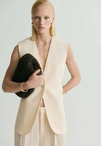 Blazer sin mangas crema con un profundo escote en V, combinado con pantalones claros. Sosteniendo un bolso texturizado de un marrón oscuro, acentuado por joyería dorada.