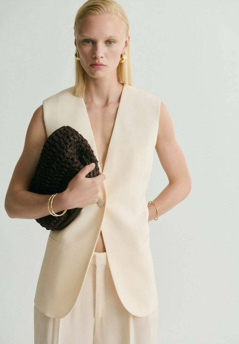 Cremfarbener ärmelloser Blazer mit tiefem V-Ausschnitt, kombiniert mit hellen Hosen. Hält eine strukturierte dunkelbraune Tasche, akzentuiert durch goldenen Schmuck.