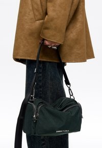 Bolsa de tela verde oscuro con dos compartimentos con cremallera, correa negra y herrajes plateados. Logo "BIMBA Y LOLA" en la parte frontal.