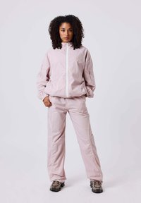 Mujer con cabello rizado vistiendo un chándal rosa claro y zapatillas de deporte con estampado, de pie con las manos en los bolsillos frente a un fondo liso.