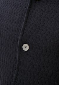 Navy textured button-up shirt featuring a single light-colored button with four holes, highlighting a woven pattern on the fabric.