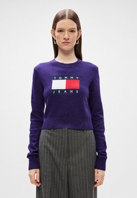 Μωβ κοντομάνικο πουλόβερ με λευκό και κόκκινο ορθογώνιο λογότυπο. Ριμπ σορτς και μανσέτες; το ύφασμα έχει ελάχιστη υφή με διακριτές πιτσιλιές.