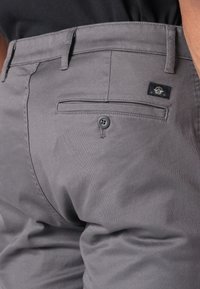 Pantalones grises con una textura suave, pasadores para cinturón, bolsillo trasero con cierre de botón y una etiqueta de marca cerca del bolsillo.