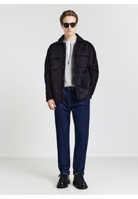 Schwarze Wildlederjacke mit Brusttaschen, grauer Pullover und dunkelblaue strukturierte Hose. Schwarze Schnürschuhe und runde Sonnenbrille runden den Look ab.