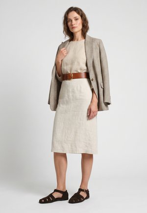 Vrouw draagt een beige jurk met een bruine riem, geruite blazer over de schouders gedrapeerd en donkerbruine sandalen, staand tegen een witte achtergrond.