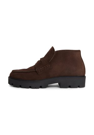 Brązowe zamszowe botki za kostkę z grubą czarną podeszwą i detalem penny loafer na białym tle.