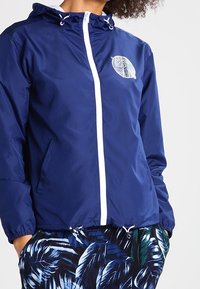 Chubasquero azul marino con cremallera blanca y logotipo, que incluye capucha y puños elásticos. Combinado con pantalones cortos estampados con hojas tropicales.
