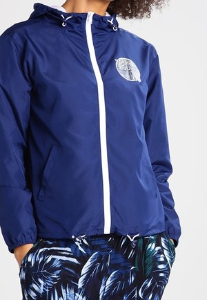 Marineblauwe windbreaker met een witte rits en logo, voorzien van een capuchon en elastische manchetten. Gecombineerd met gebloemde shorts met tropische bladeren.