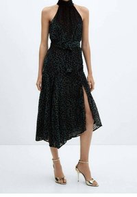 Schwarzes Halfterkleid mit einem gepunkteten Muster, das einen seitlichen Schlitz und ein gebundenes Taillendetail aufweist. Getragen mit goldenen Riemchen-High Heels.