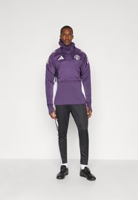 Lilla kvart-zip sportsjakke med hætte, hvide sidepaneler og et logo. Sammen med sorte tapered sweatpants og sorte sko.