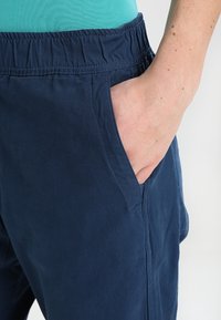 Granatowe spodnie z elastycznym pasem, wyposażone w boczną kieszeń, wykonane z gładkiego materiału. Widok z bliska skupiający się na kieszeni.