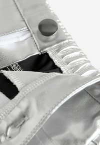 Silberne Metallic-Stoff mit glänzender Textur, ausgestattet mit einem schwarzen Knopf und sichtbaren Nähten, die einen teilweise geöffneten Reißverschluss zeigt.
