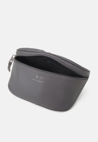 Szara skórzana saszetka o zaokrąglonym kształcie, wyposażona w górny zamek błyskawiczny oraz wewnętrzną kieszonkę. Na przedniej powierzchni widoczne logo marki.