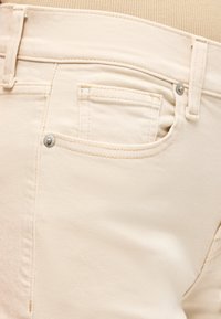 Κοντινή λήψη σε μπεζ τζιν από denim που διαθέτουν μπροστινή τσέπη με μικρή τσέπη για κέρματα, θηλιές για ζώνη και μεταλλικά καρφιά στη μέση και στην άκρη της τσέπης.