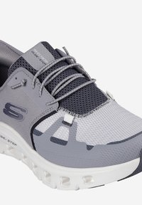 Zapatilla deportiva gris que presenta materiales de malla y sintéticos, un acabado texturizado y un diseño con cordones con acentos recortados.