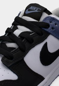 Κοντινή λήψη ενός μαύρου, άσπρου και μπλε αθλητικού παπουτσιού Nike με μαύρα κορδόνια και το λογότυπο της Nike στο πλάι και στη γλώσσα του παπουτσιού.