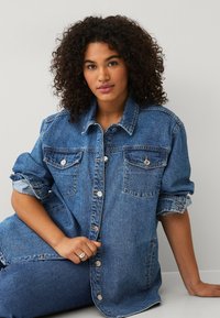 Giacca di denim di blu medio, dotata di due tasche sul petto, bottoni in metallo argentato e maniche arrotolate con un design a vestibilità rilassata.