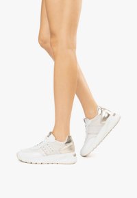 Baskets de sport blanches avec des accents métalliques, des détails en mesh et une semelle rembourrée, mettant en avant un bout rond et un talon contrastant.