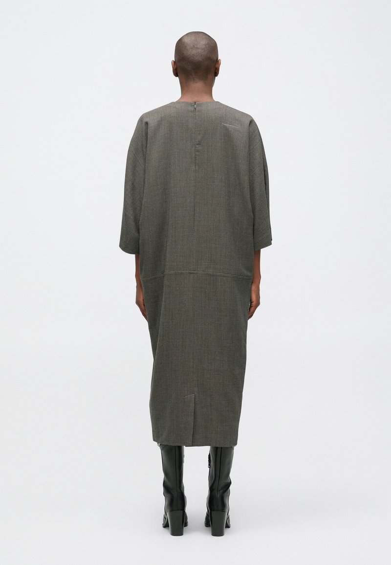 Robe grise longue et ample avec une texture subtile, des manches trois-quarts, une fermeture au cou à l'arrière et une fente haute. Associée à des bottes noires montantes.