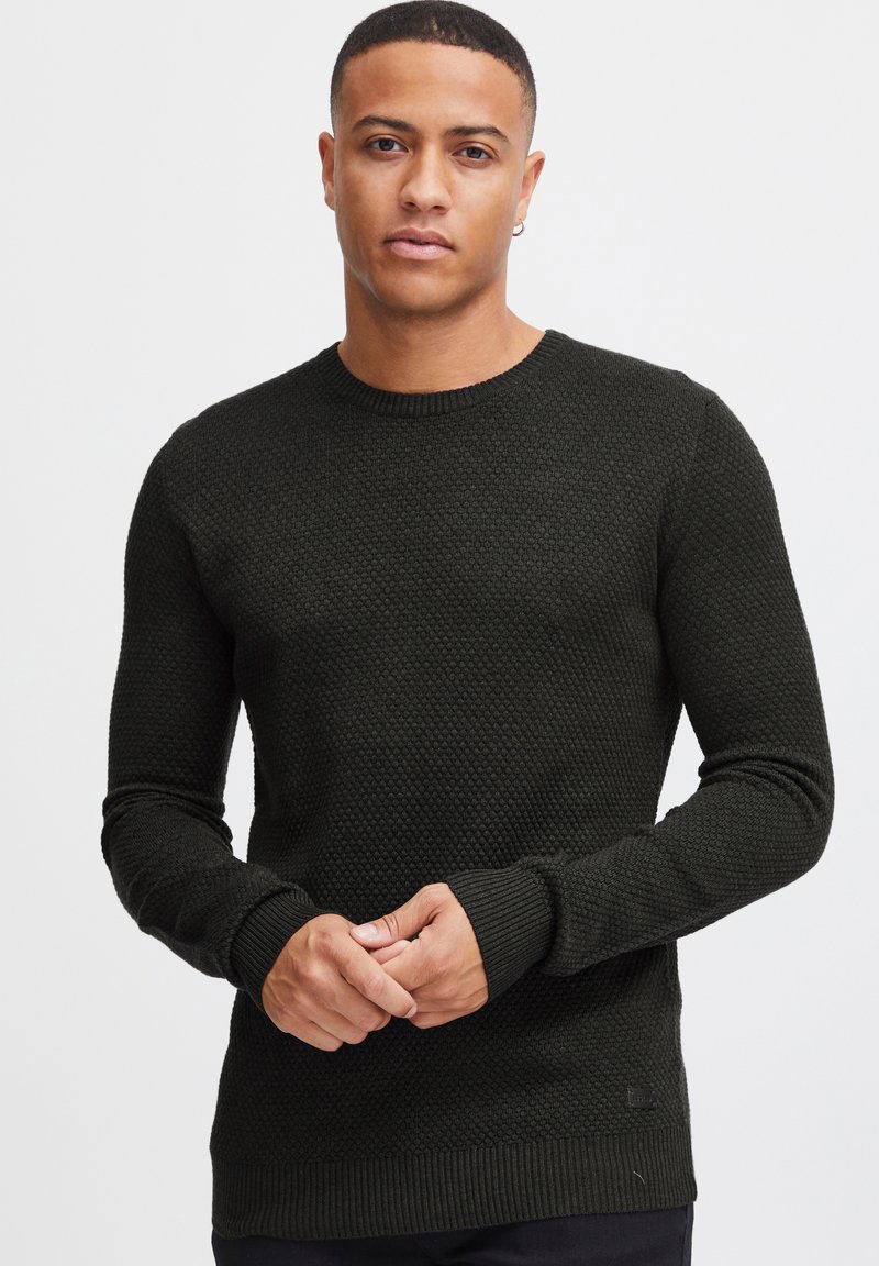 Maglione lavorato a maglia verde scuro con un motivo testurizzato, scollo rotondo, polsini e orlo a costine. Il tessuto appare morbido e leggermente elastico.