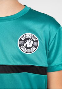Teal athletic shirt with a black horizontal stripe. Features a round embroidered logo with a gorilla design and the text "GORILLA WEAR."